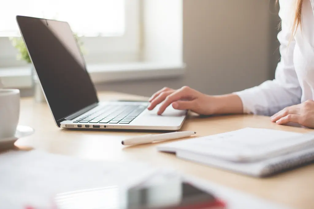 A person typing on a laptop at a desk, with a notebook, a pen and a cup beside it.